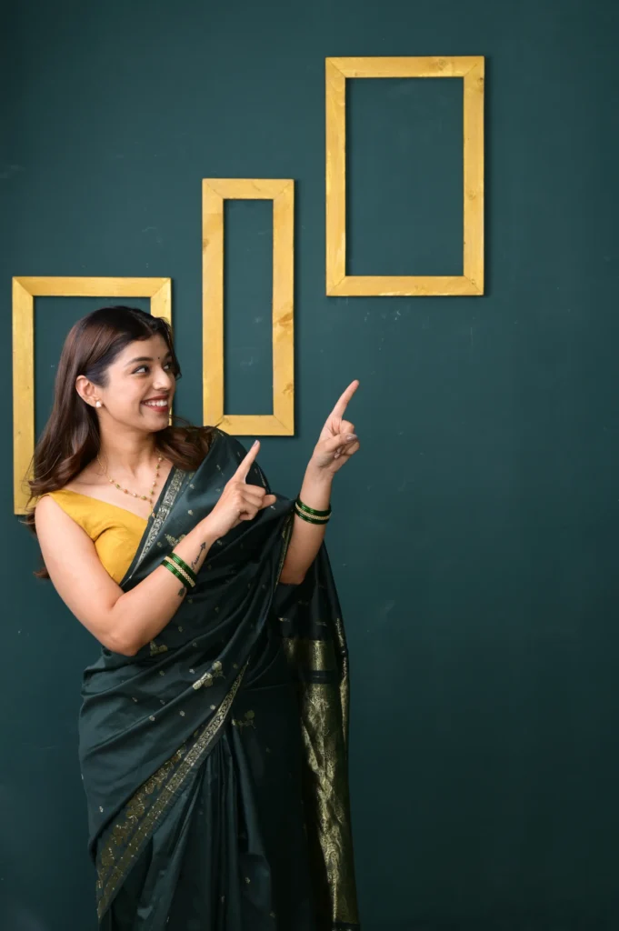 Woman in elegant green saree with golden border smiling and pointing towards empty wooden frames on dark green wall background.